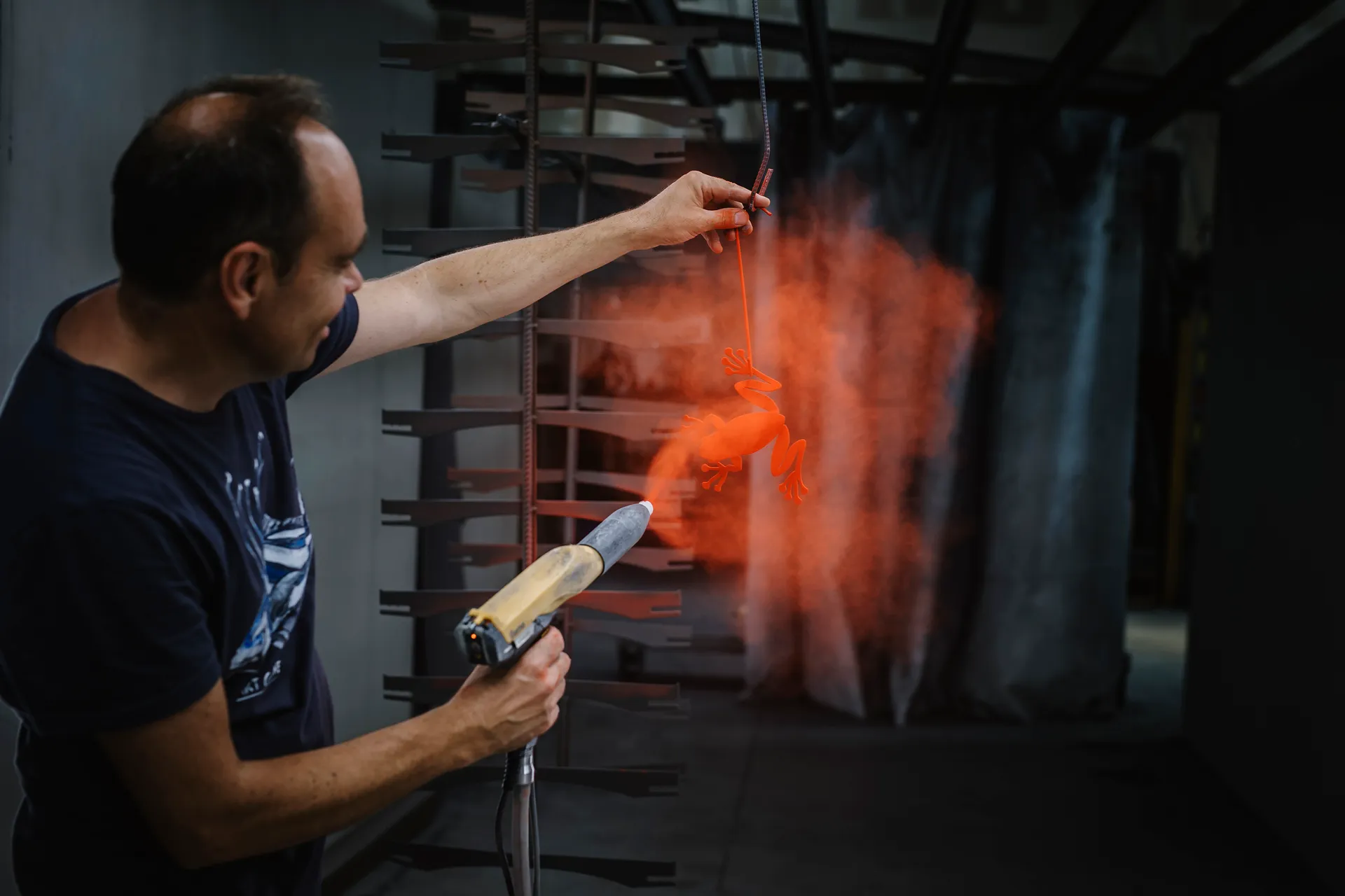 l'artiste projetant un nuage de peinture orange à l'aide d'un pistolet sur une grenouille de métal